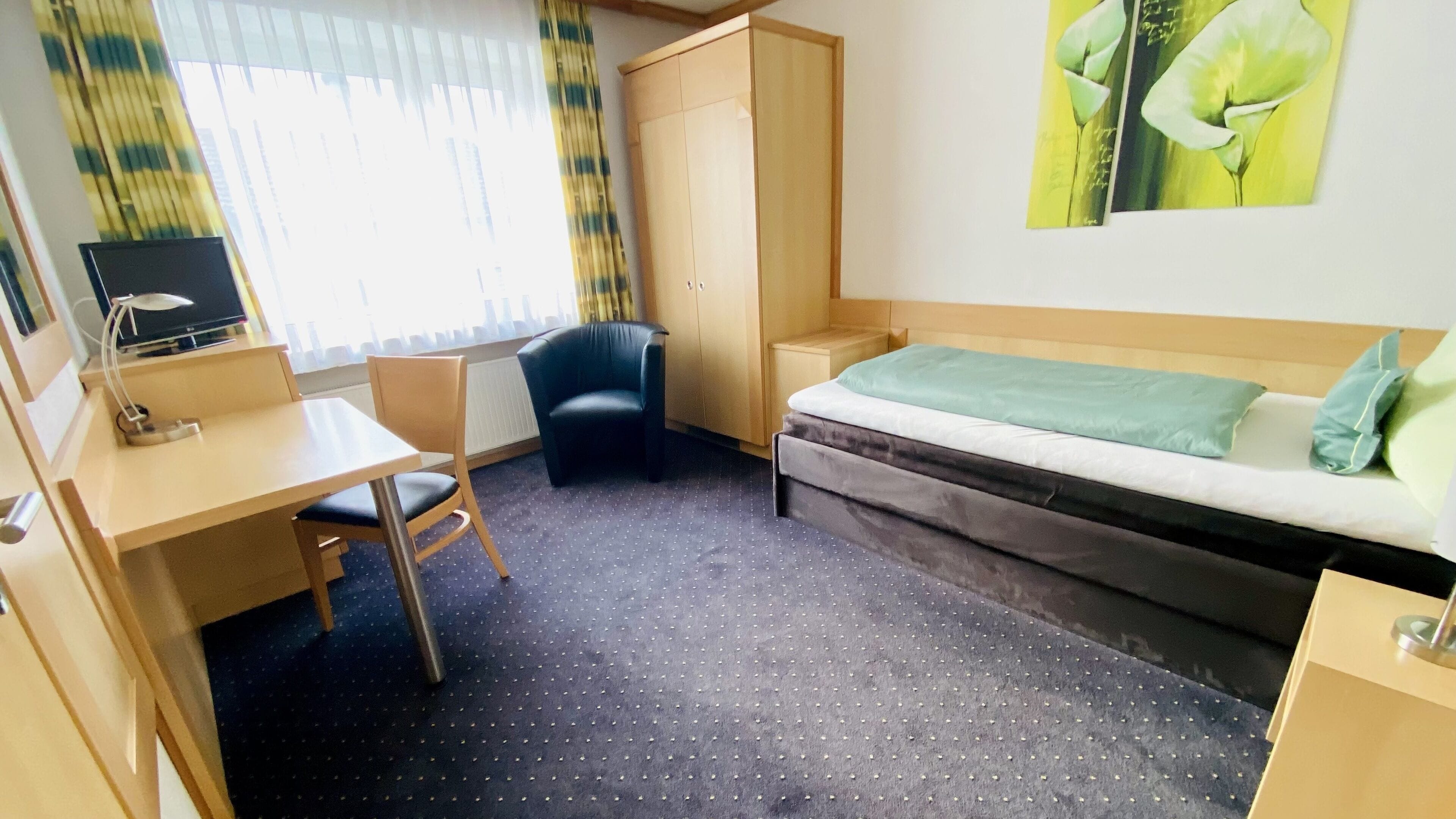 Ein Hotelzimmer mit Schreibtisch, Stuhl, Kleiderschrank, Bett und einem Gemälde an der Wand.