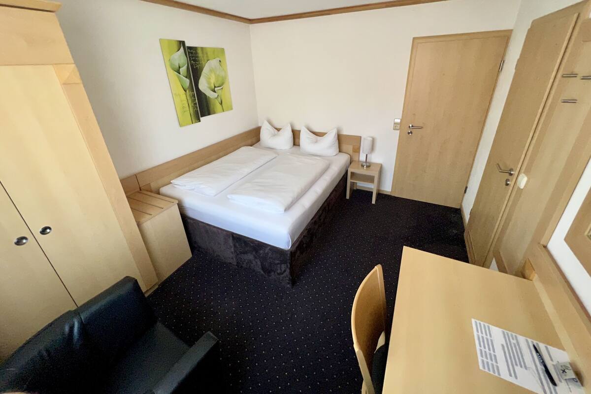 Ein Hotelzimmer mit Bett, einem kleinen Tisch, einem Sessel, einem Nachttisch, einem Kleiderschrank und einem Wandbild.