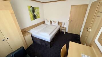 Una habitación de hotel con cama, una mesita, una silla, una mesita de noche, un armario y un cuadro en la pared.