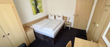 A hotel room with a bed, a small table, a chair, a nightstand, a wardrobe, and a wall with a framed picture.