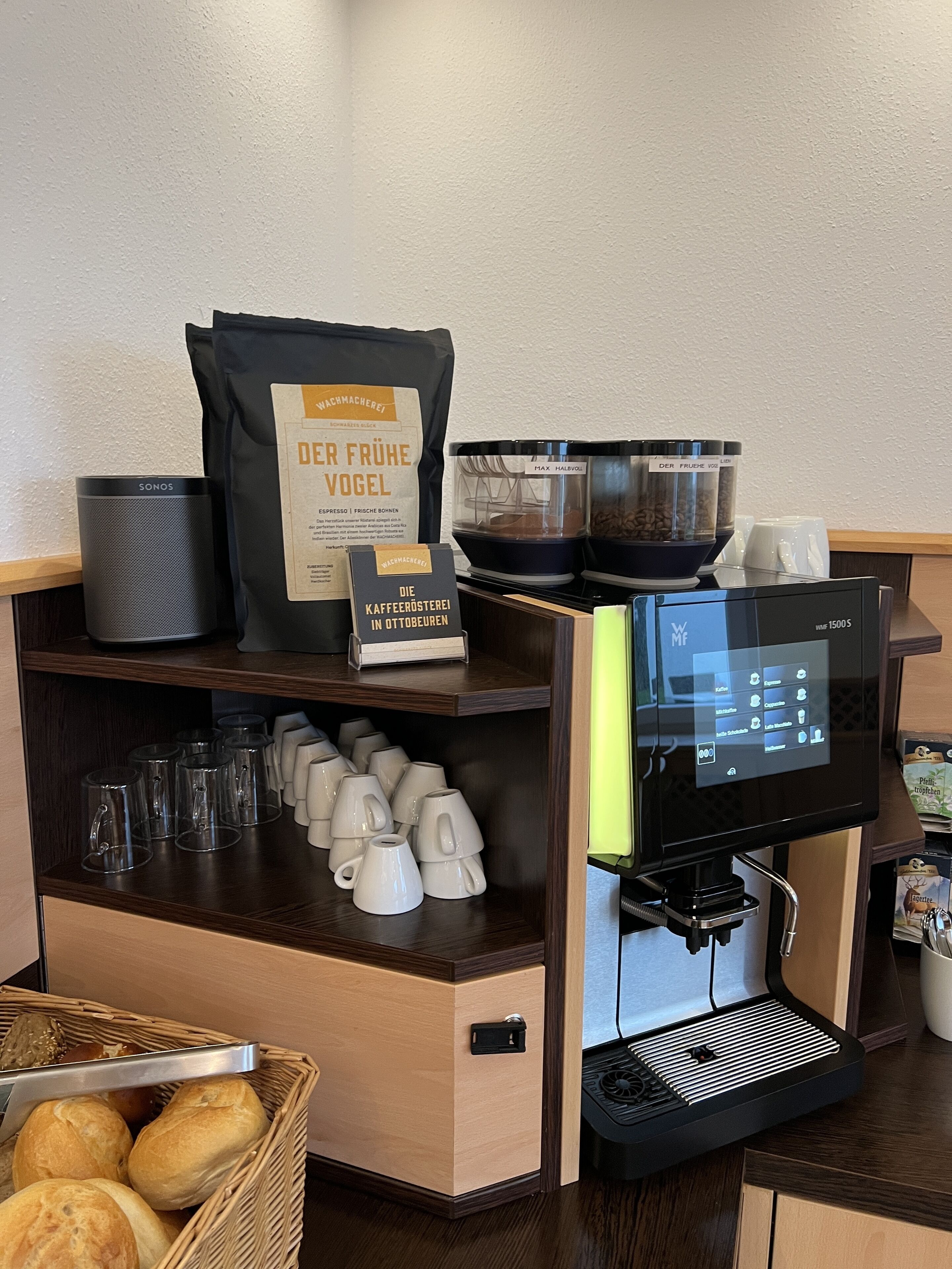 Una estación de café con una máquina de café, tazas, un paquete de granos de café y un letrero que indica que el café es de Ottobeuren.