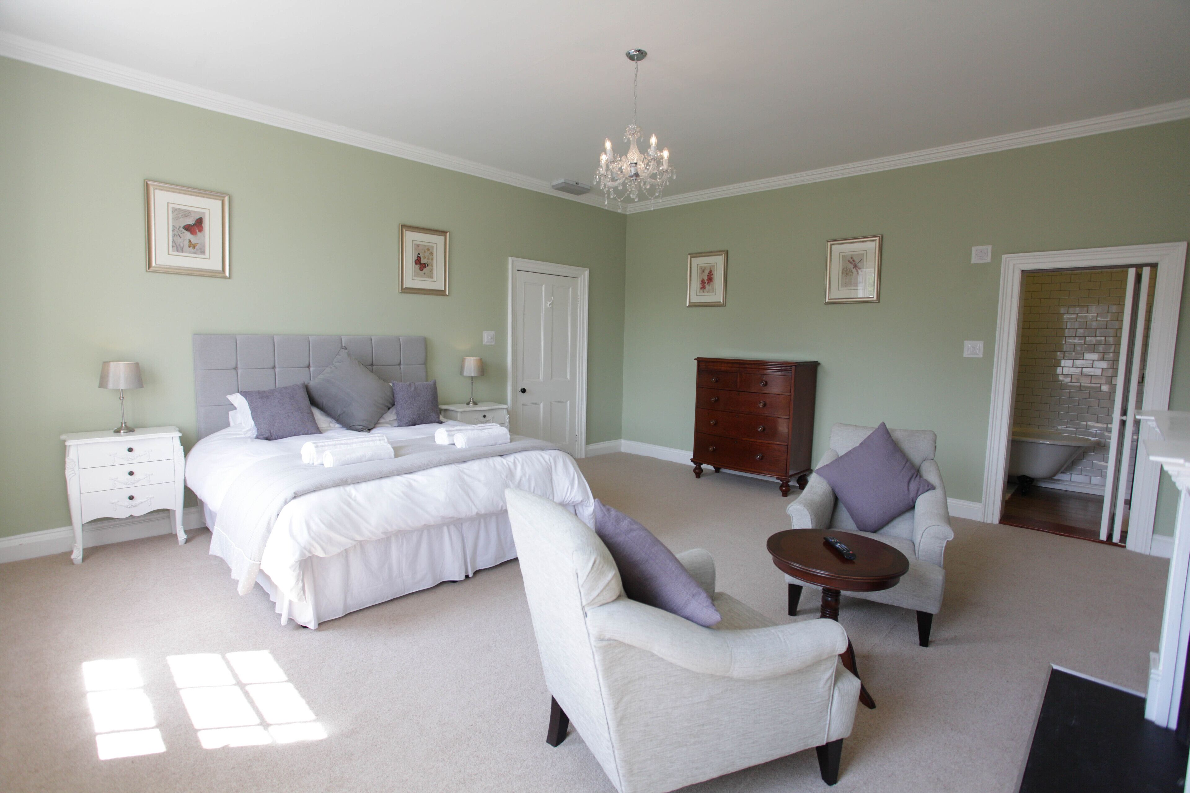 A spacious bedroom with a large bed, a dresser, two armchairs, a small table, and a chandelier.