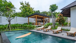 Una piscina con sillones de descanso, una terraza de madera y una zona de estar cubierta.