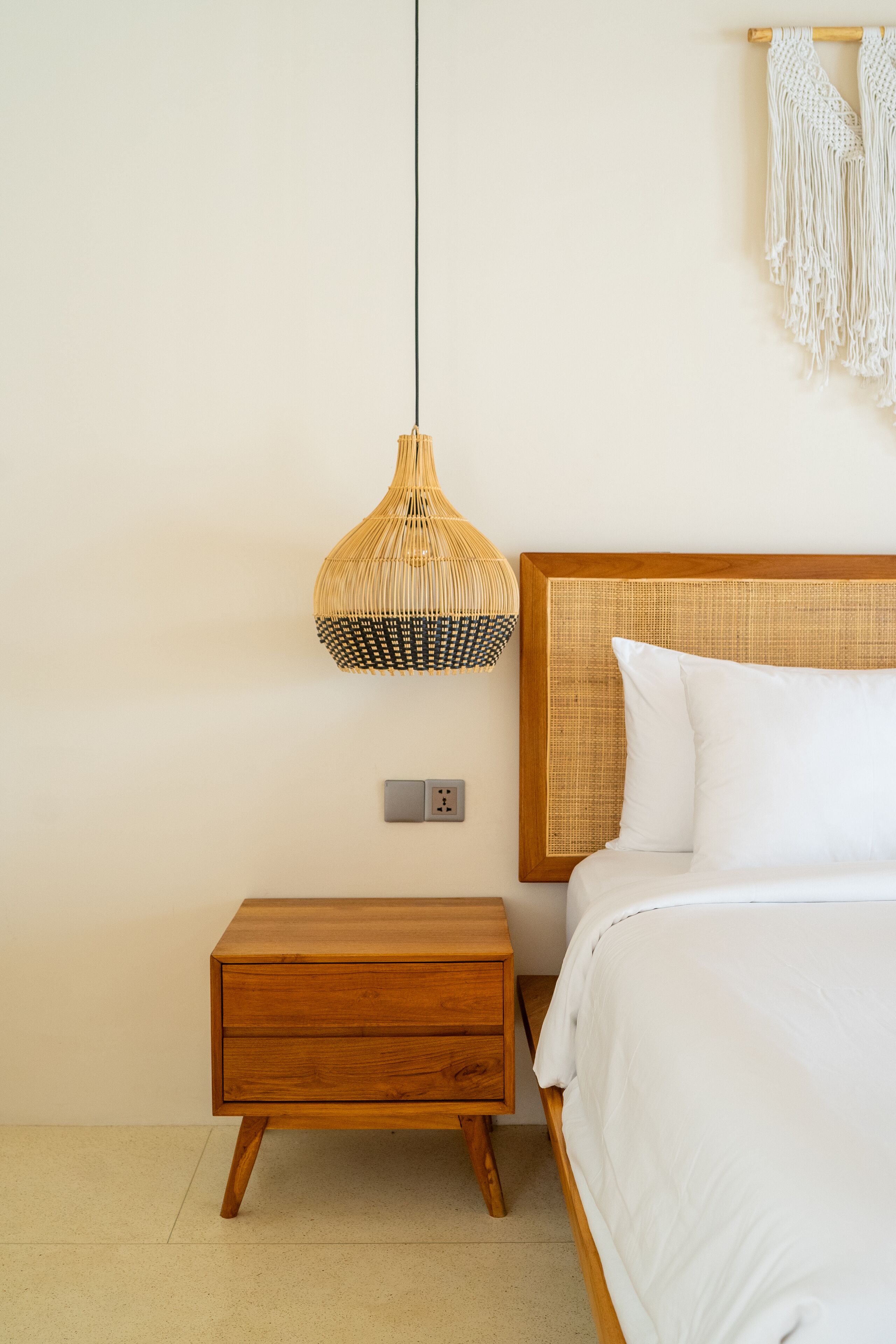 Ein Schlafzimmer mit einem Nachttisch aus Holz, einem Bett mit weißer Bettwäsche, einer gewebten Pendelleuchte und einem Wandbehang aus Makramee.