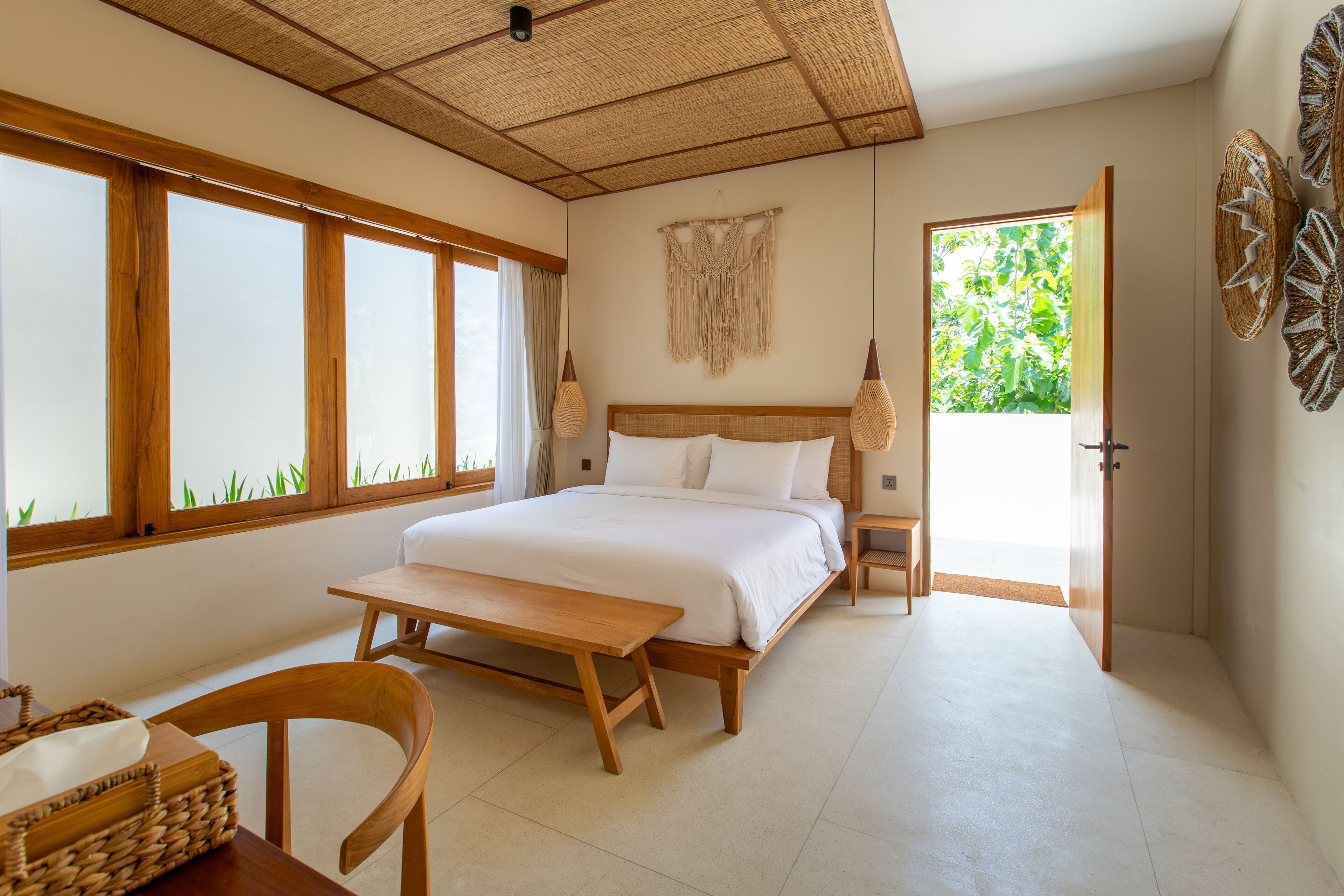 Un dormitorio con una cama, un banco de madera, una silla, una mesita y una ventana con vistas a la vegetación.