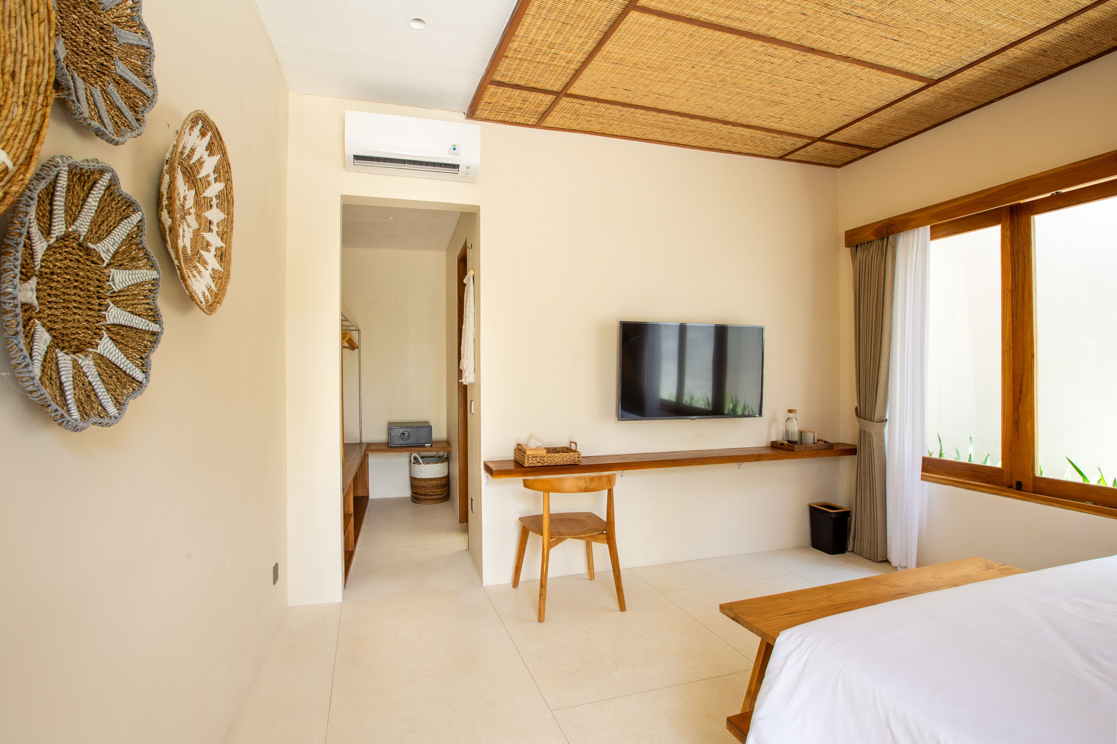 A modern hotel room with a large bed, a TV mounted on the wall, a wooden desk, and a view of the outdoors through a large window.