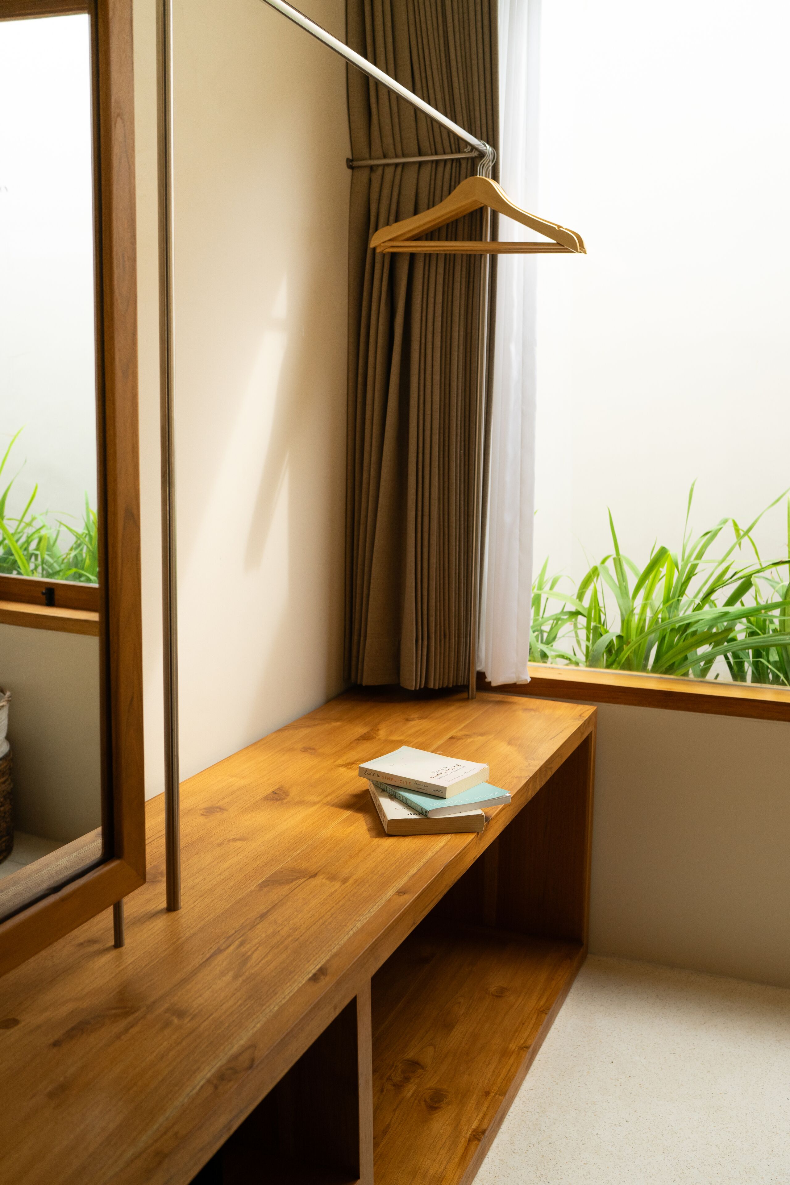 A corner room with a wooden bench, a mirror, a wooden hanger, and a window with curtains.