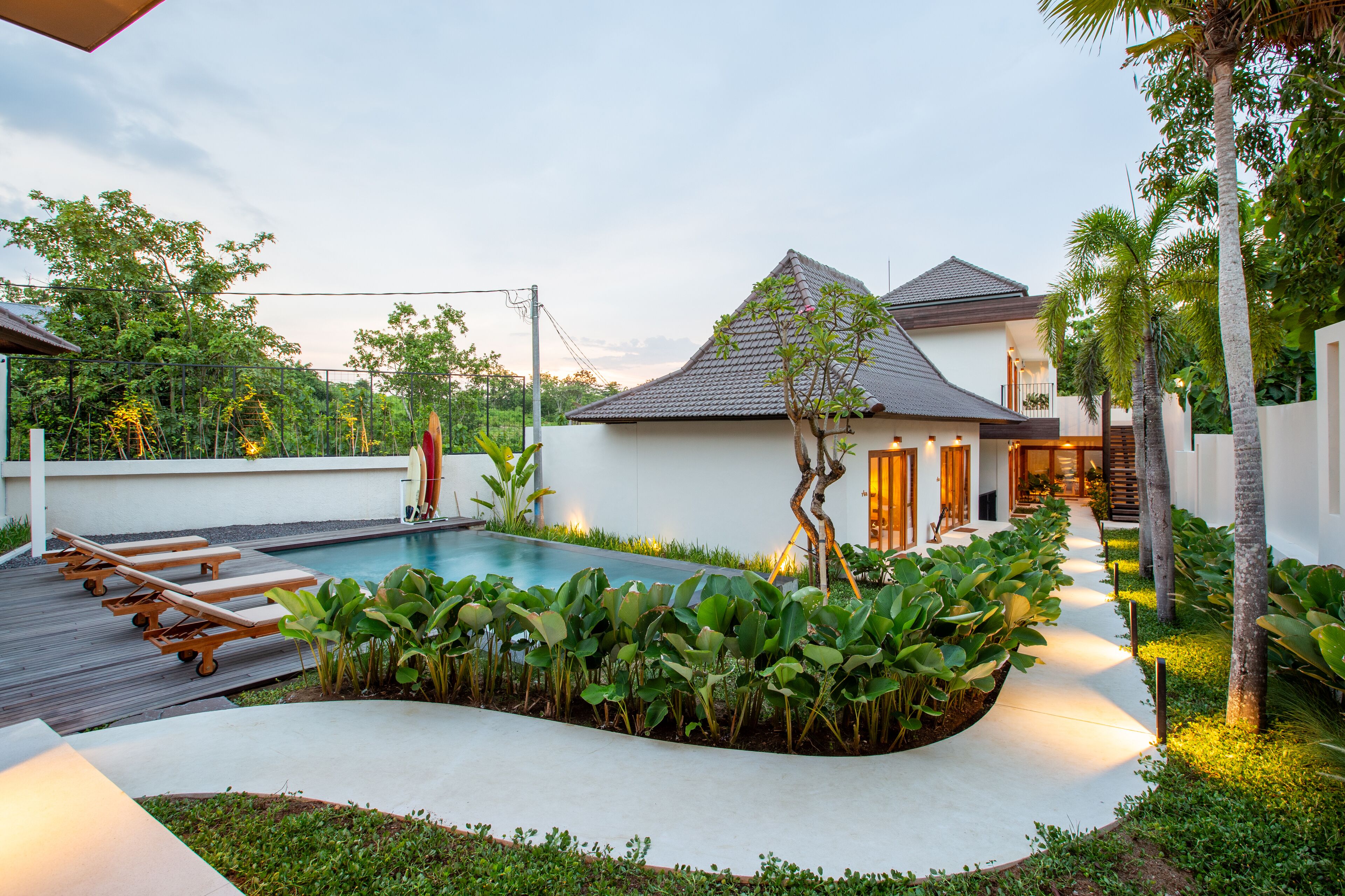 A pool area with lounge chairs, a small house with a tiled roof, and a pathway lined with plants.