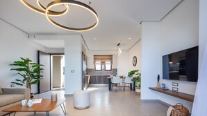 A modern living room with a sofa, coffee table, and a TV. There is a kitchen in the background with a dining area. - Riviera Vista Luxury Homes, Mousela 2broom suite with sea view and shared pool (Dramia)