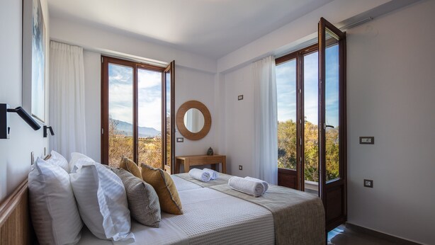 A bedroom with a bed, pillows, a bedside table, a mirror, and large windows offering a view of the outdoors. - Riviera Vista Luxury Homes, Mousela 2broom suite with sea view and shared pool (Dramia)