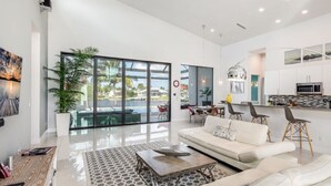 A modern living room with a large glass door leading to a pool, a white sofa, a coffee table, and a kitchen area with a microwave. - Villa Rapa Nui Island (Cape Coral)