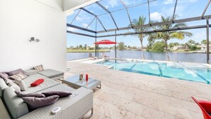 A modern outdoor pool area with a glass enclosure, a seating area with cushions, and a view of a body of water. - Villa Rapa Nui Island (Cape Coral)