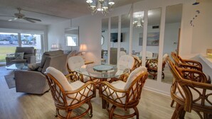 A spacious living room with a glass coffee table surrounded by wicker chairs, a ceiling fan, and a large window offering a view of the outdoors.