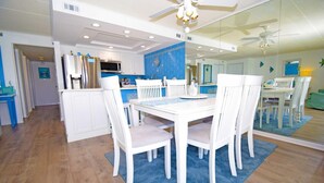 A modern kitchen with white cabinetry, a stainless steel refrigerator, and a microwave. There is a dining area with a white table and chairs, and a blue rug on the floor.