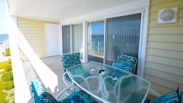 A balcony with a glass table and chairs, a sliding glass door, and a view of the ocean.