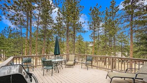 A wooden deck with outdoor furniture, including a table and chairs, surrounded by tall pine trees. - Hidden Pines -- Ev#3034 2 Bedroom Home by RedAwning (Estes Park)