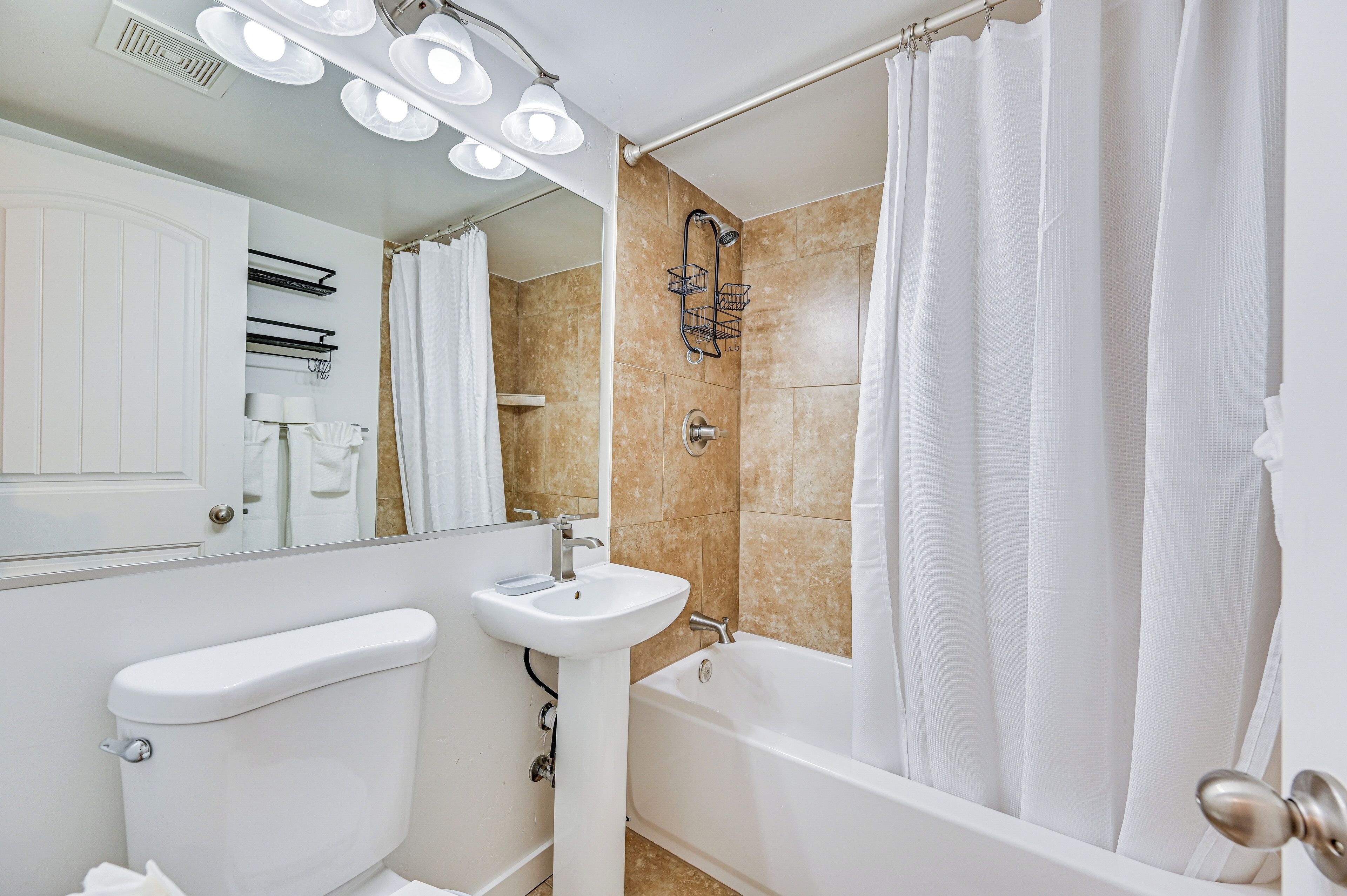 Ein Badezimmer mit weißem Waschbecken, WC und Badewanne, einer Duschvorhang sowie einem Spiegel mit Beleuchtung darüber.