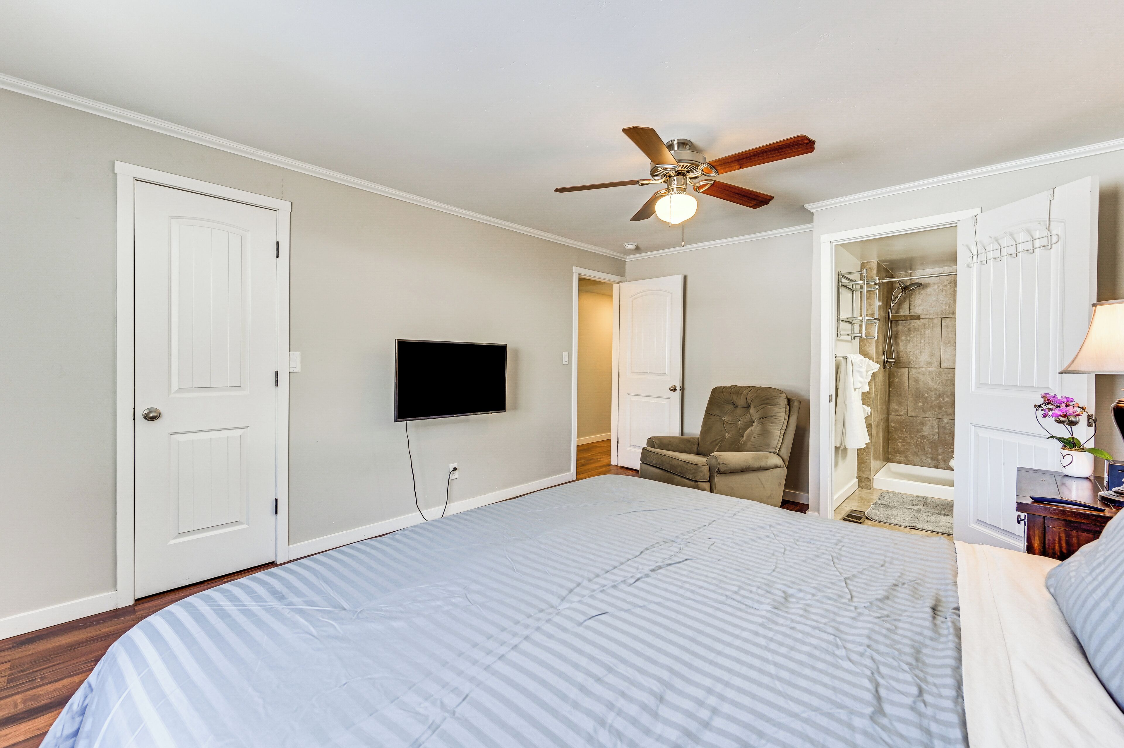 Une chambre avec un lit, un ventilateur de plafond, un téléviseur fixé au mur et un espace douche visible à travers une porte ouverte.