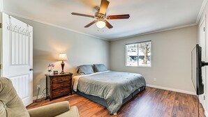 Quarto com cama, mesa de cabeceira, abajur, ventoinha no teto, janela e piso de madeira.
