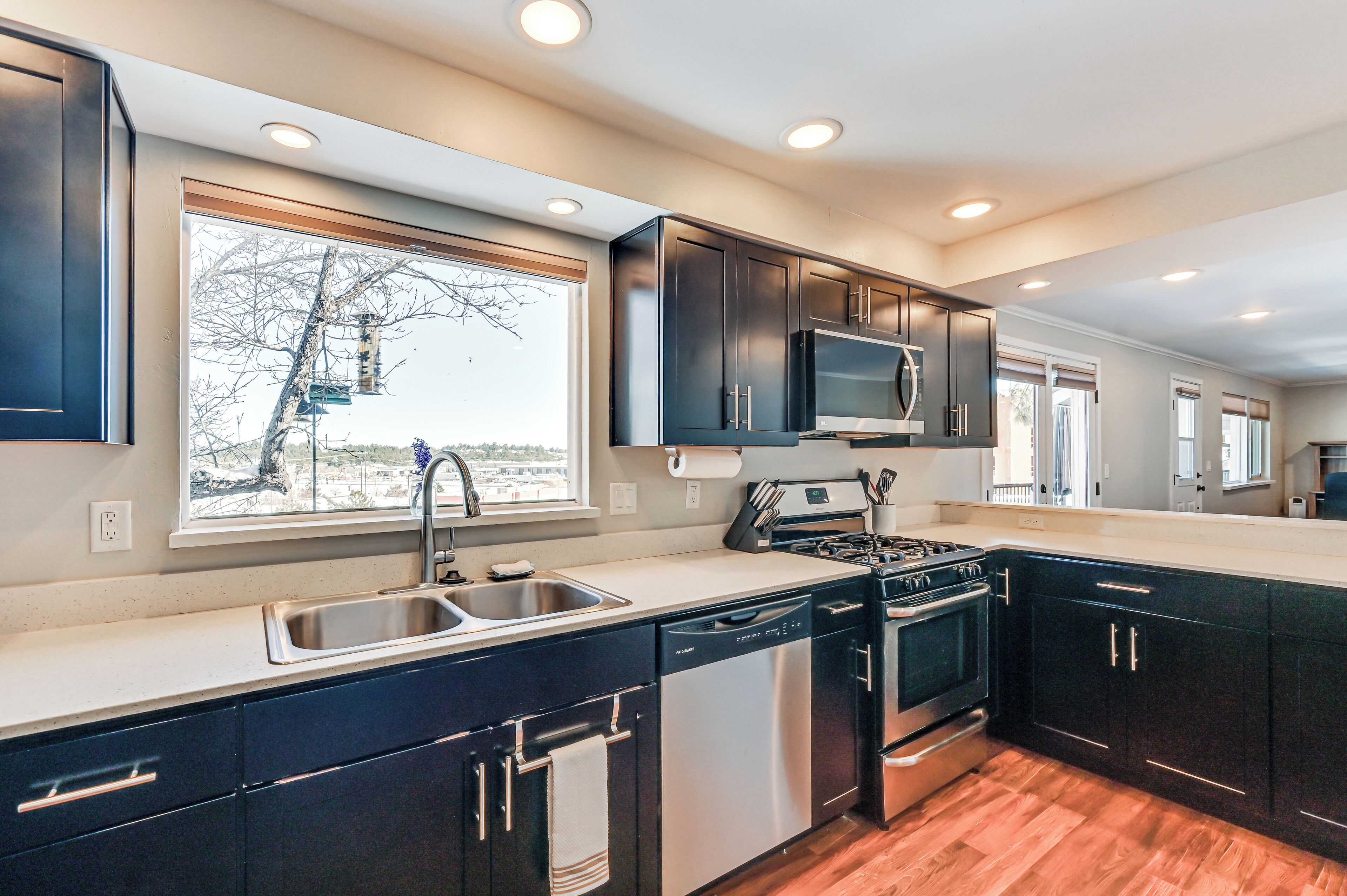 A modern kitchen with dark cabinets, stainless steel appliances, a large window, and a view of a tree outside.
