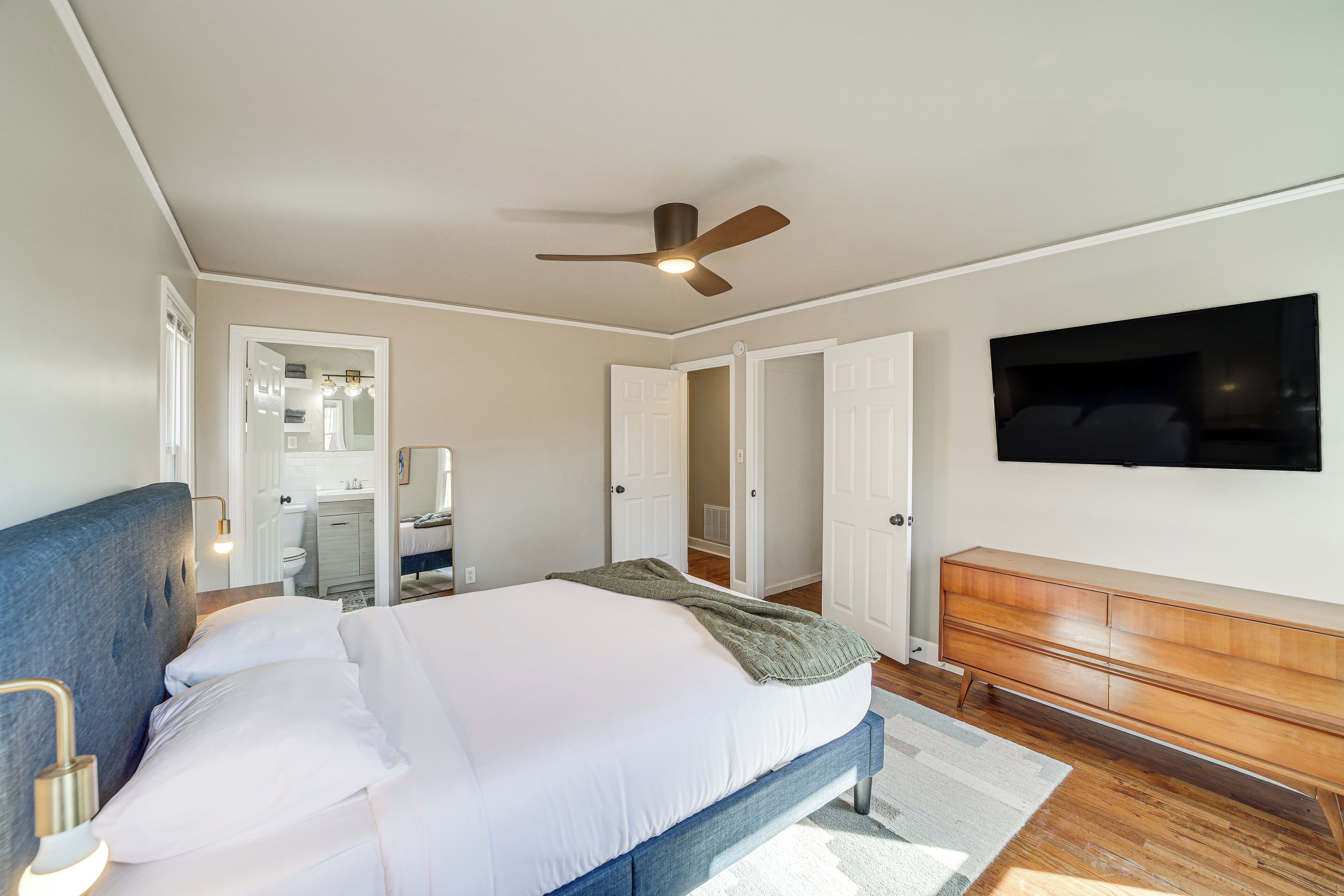 Un dormitorio con una cama, un ventilador de techo, un televisor montado en la pared y un aparador de madera.