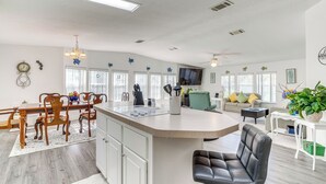 A modern kitchen with a central island, dining area with wooden chairs, a sofa, and a television in the living room.
