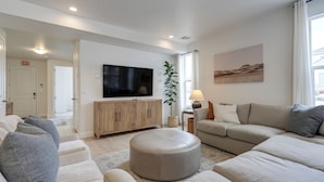 Uma sala de estar moderna com sofá bege, um banquinho redondo, uma TV de tela plana fixada em um móvel de madeira e uma janela grande com cortinas.