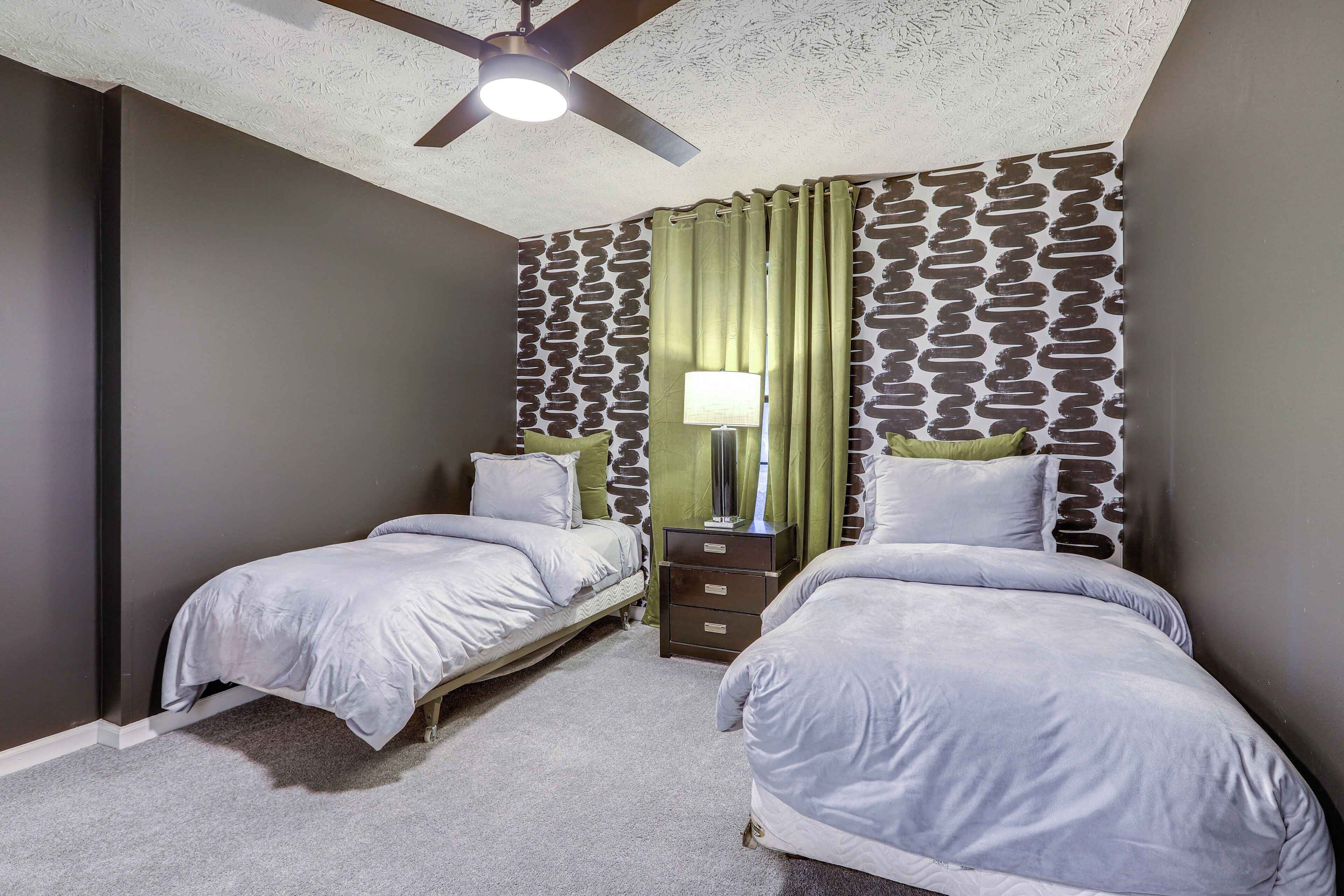 A bedroom with two beds, a ceiling fan, a nightstand with a lamp, and patterned wallpaper.