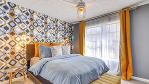 Une chambre avec un mur à motifs, un lit avec une literie bleue, une table de chevet et une fenêtre avec des rideaux jaunes.