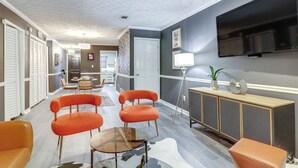 Un salon moderne avec une grande télévision, des fauteuils orange, une table basse en verre et un meuble en bois.