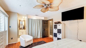 Une chambre à coucher avec un lit, un fauteuil blanc, un ventilateur de plafond, une télévision et un dressing.