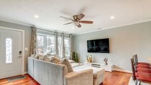 A living room with a grey sofa, a wooden floor, a ceiling fan, a TV mounted on the wall, and a door with a decorative glass panel. - Spacious Home w/ Tranquil Patio ~ 29 Mi to DC! (Severn)