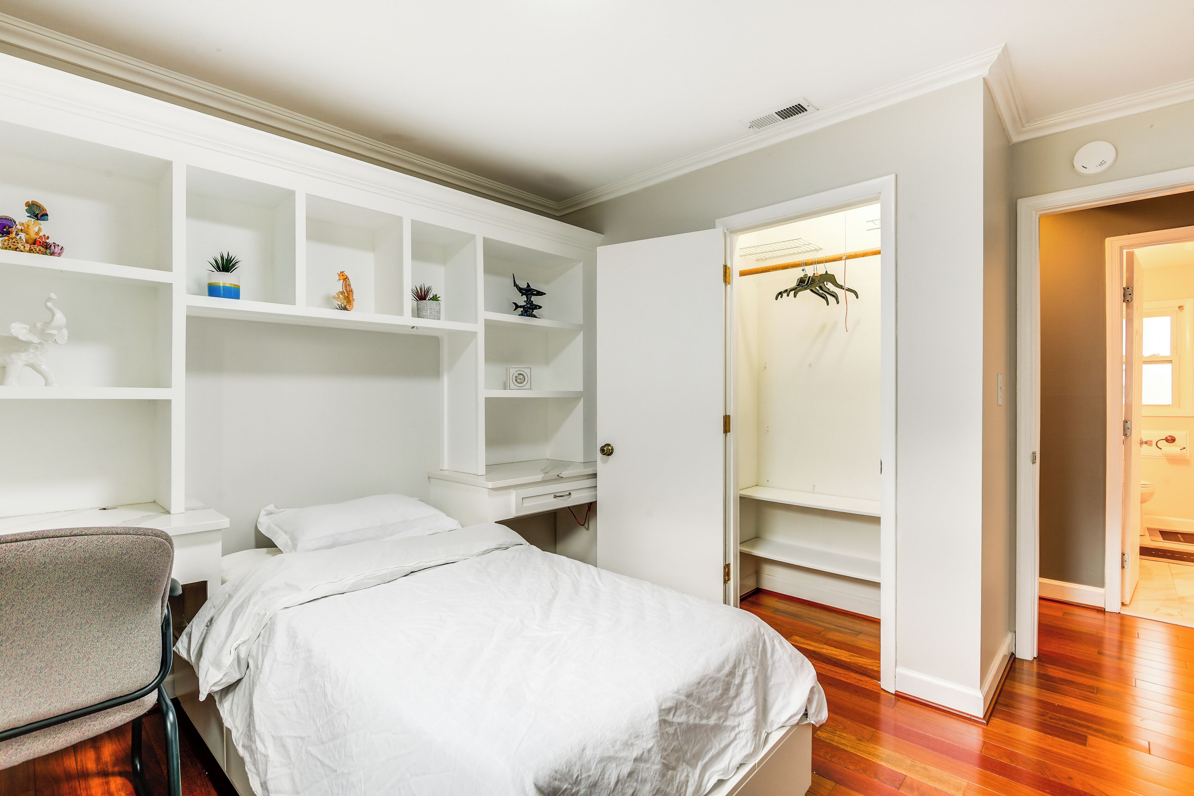 Une chambre à coucher avec un lit, un bureau et une chaise. On y trouve des étagères ornées d’objets décoratifs et une armoire contenant des vêtements suspendus.