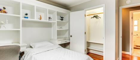 Une chambre à coucher avec un lit, un bureau et une chaise. On y trouve des étagères ornées d’objets décoratifs et une armoire contenant des vêtements suspendus.