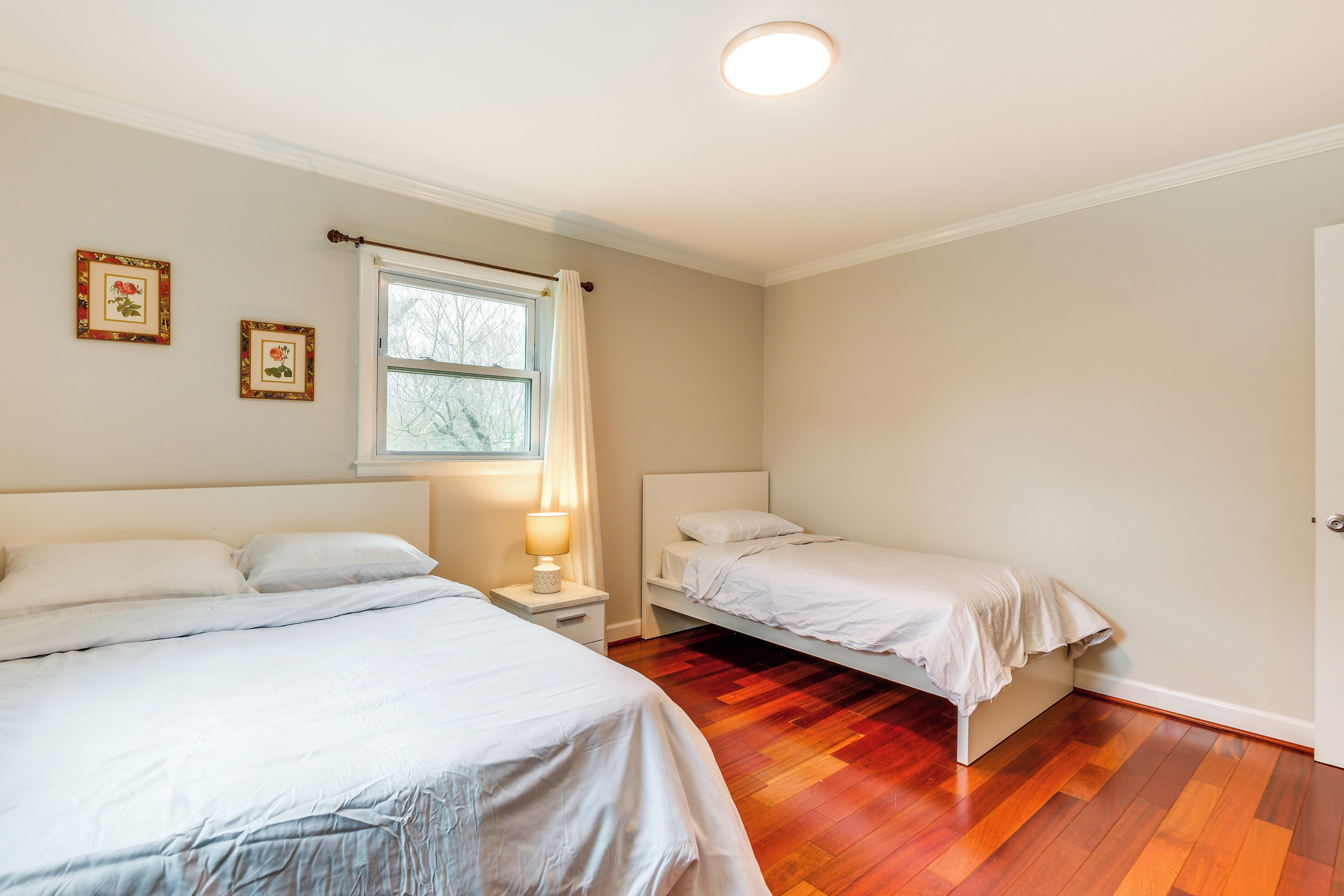 Un dormitorio con dos camas, una ventana con cortinas, una mesita de noche con lámpara y suelo de madera.
