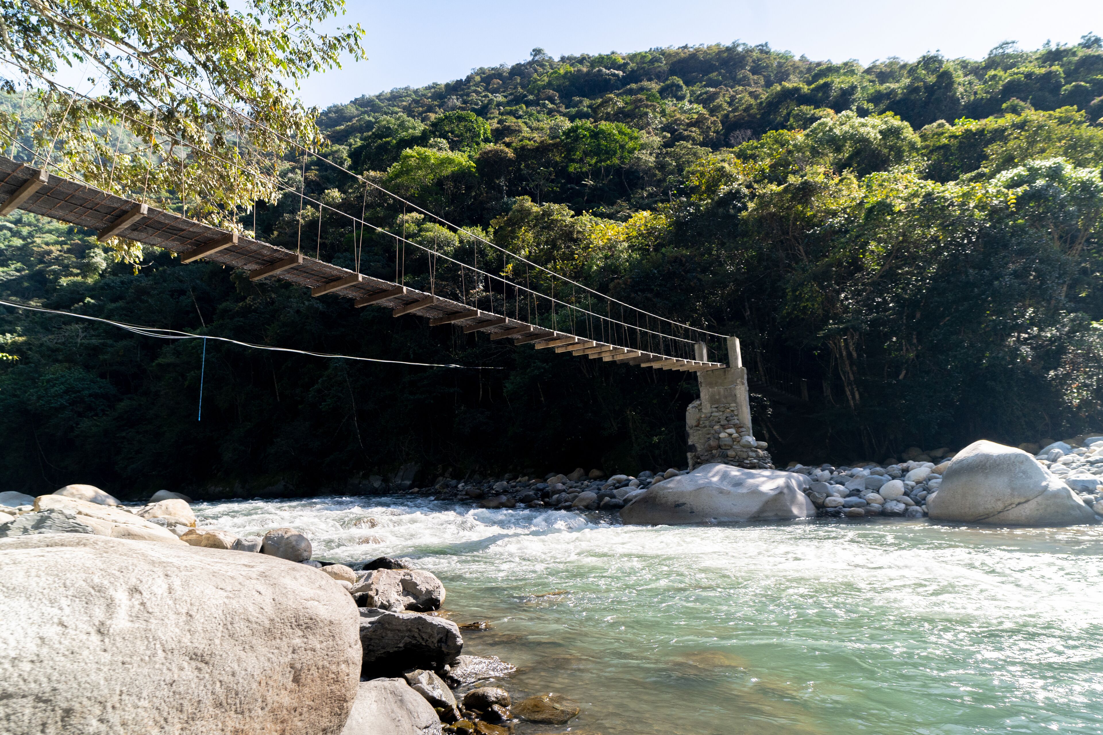 Un puente colgante de madera sobre un río con rocas y un páramo boscoso al fondo.
