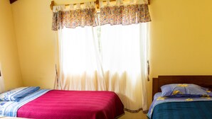 A room with two beds, each with a distinct bedspread. The room has a sloped ceiling with wooden beams, a window with a floral curtain, and a tiled floor.