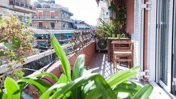 A balcony with a wooden chair, potted plants, and a view of the sea and buildings. - Classical Beauty 3Bd Apt in Nea Filothei (Athens)