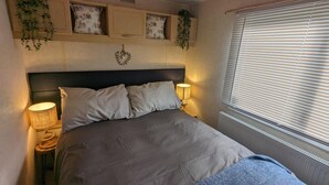 A bedroom with a bed, two bedside lamps, a window with blinds, and wall-mounted shelves with decorative items. - Sandy Paws Dog Friendly - Anderby Creek (Skegness)