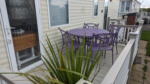 A mobile home with a patio area featuring a table and chairs, surrounded by a white fence and a large plant. - Sandy Paws Dog Friendly - Anderby Creek (Skegness)