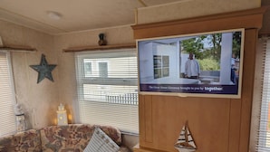 A cozy living room with a television mounted on a wooden wall, a patterned sofa, and a small sailboat decoration. - Sandy Paws Dog Friendly - Anderby Creek (Skegness)