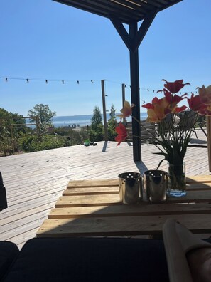 Um deck de madeira com vista para o mar, uma pérgula, luzes de corda e uma mesa com copos de metal e um vaso de flores.