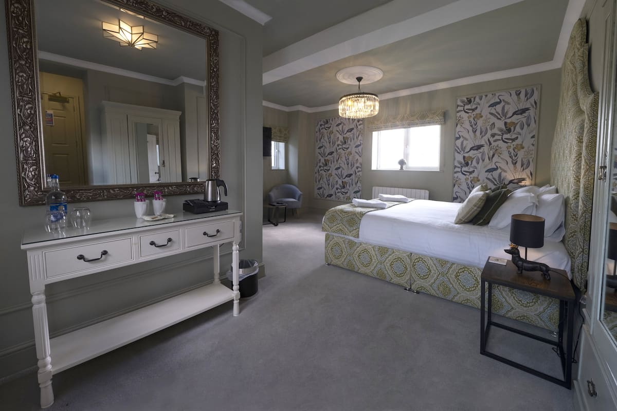 A bedroom with a large mirror, a bed with patterned bedding, a bedside table with a lamp, and a dresser with drawers.