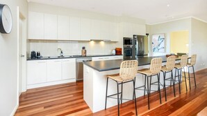 A modern kitchen with white cabinets, a central island with bar stools, stainless steel appliances, and a wooden floor. - 113 Barrage Rd Goolwa South (Goolwa South)
