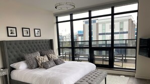 Ein modernes Schlafzimmer mit einem großen Bett, einer Bank und Blick auf Gebäude durch große Fenster.