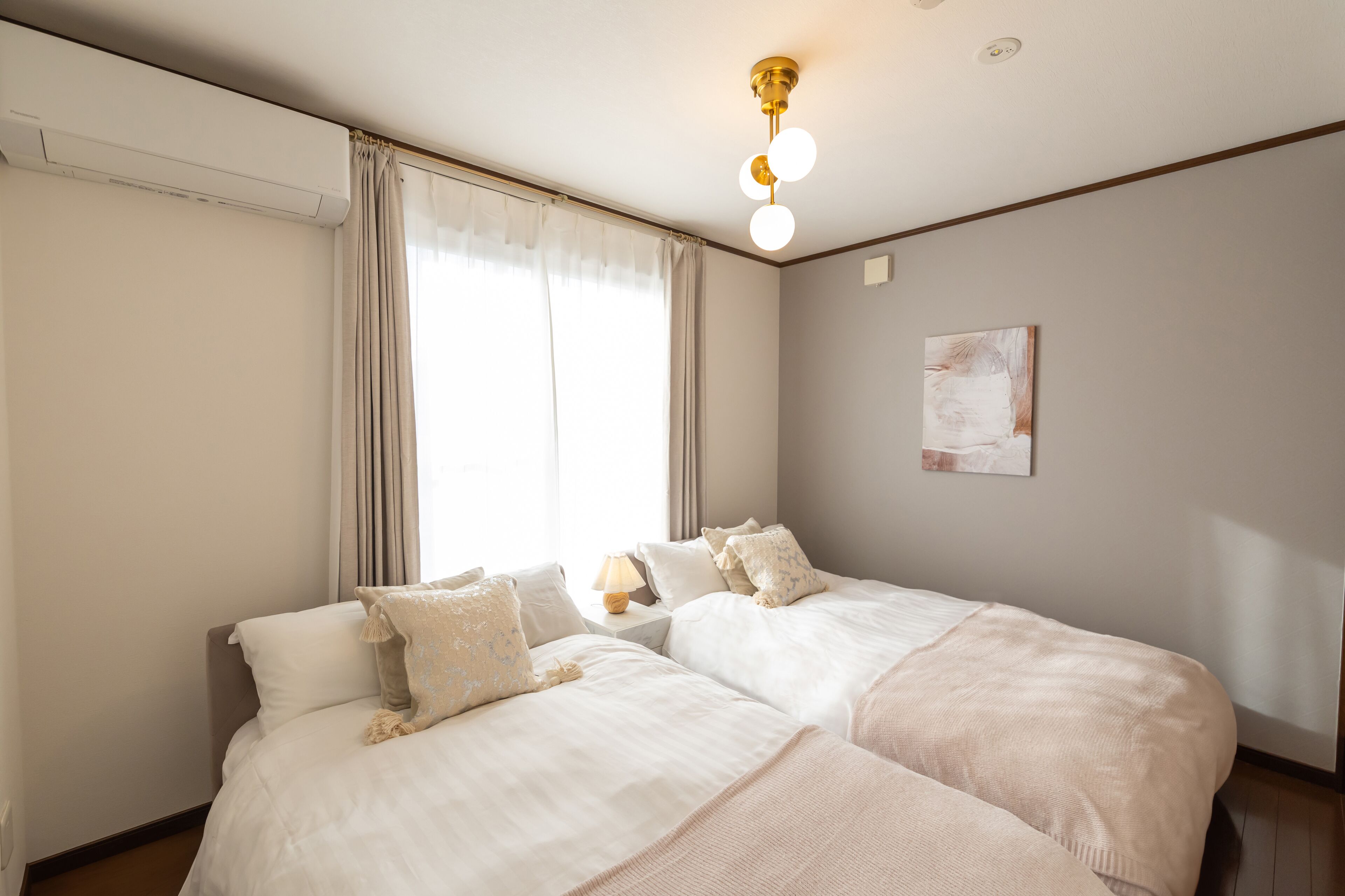 Une chambre d’hôtel avec deux lits, un luminaire au plafond, une fenêtre avec des rideaux et une œuvre d’art accrochée au mur.