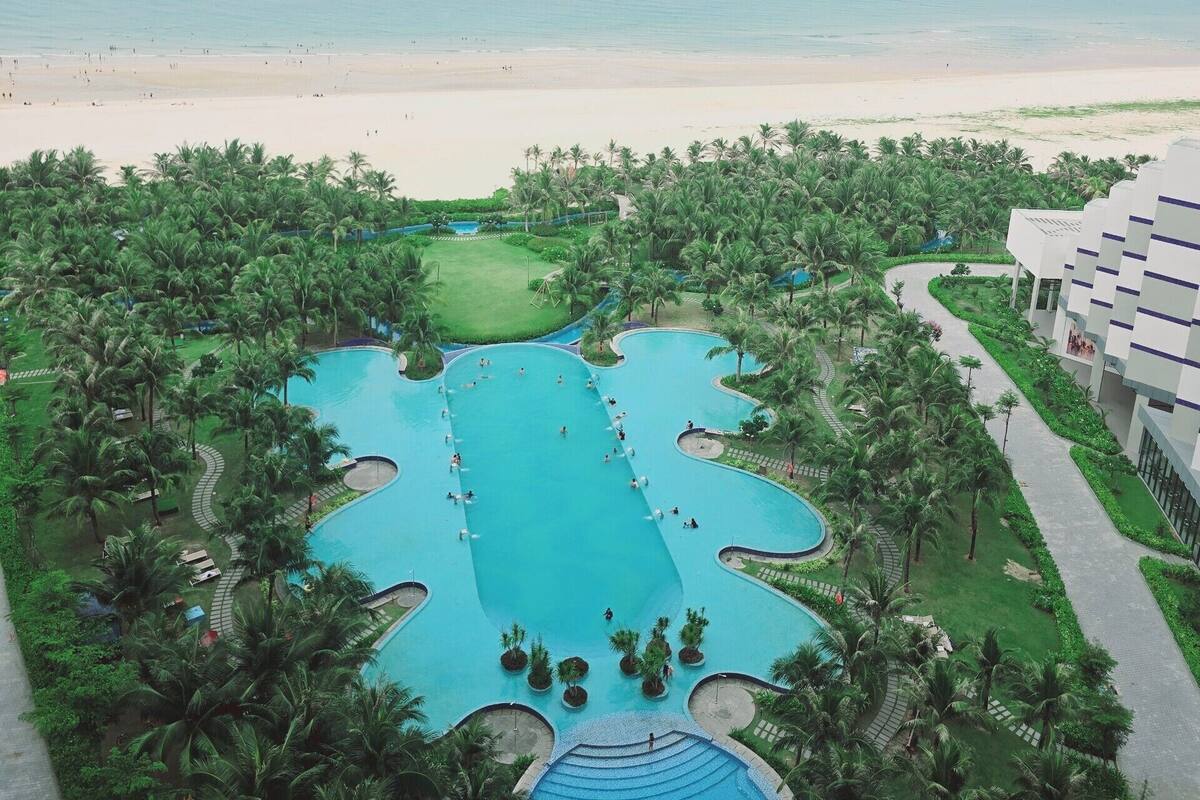 A resort with a large central pool surrounded by palm trees, a beach, and a view of the ocean.