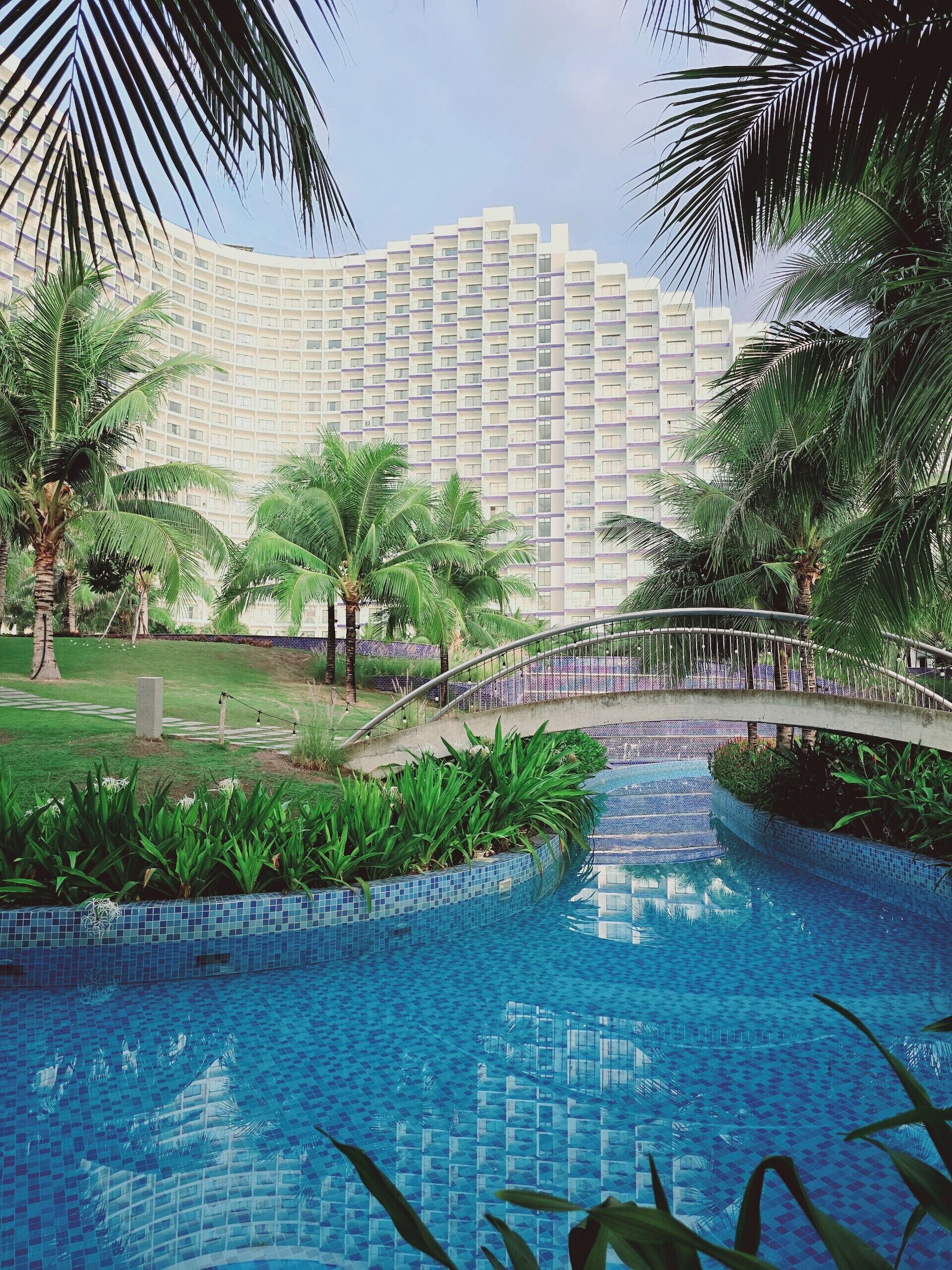 A swimming pool with a tiled edge, surrounded by greenery and palm trees, with a bridge over it leading to a high-rise building in the background.