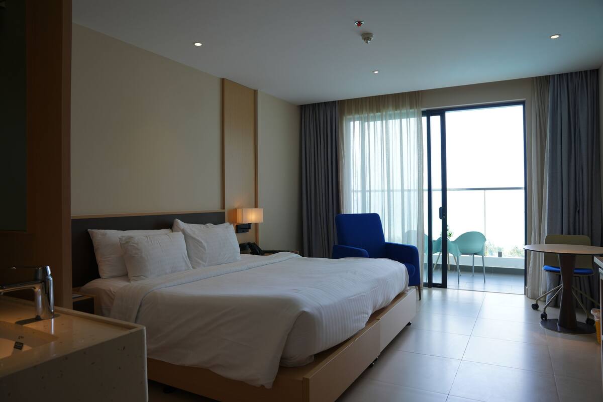 A hotel room with a large bed, a desk, a chair, and a balcony with a view.