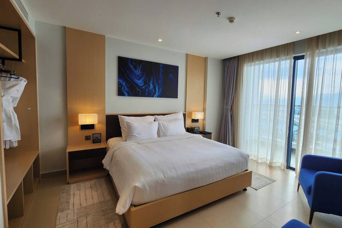 A modern hotel room with a large bed, two bedside tables with lamps, a large window with curtains, and a blue chair.
